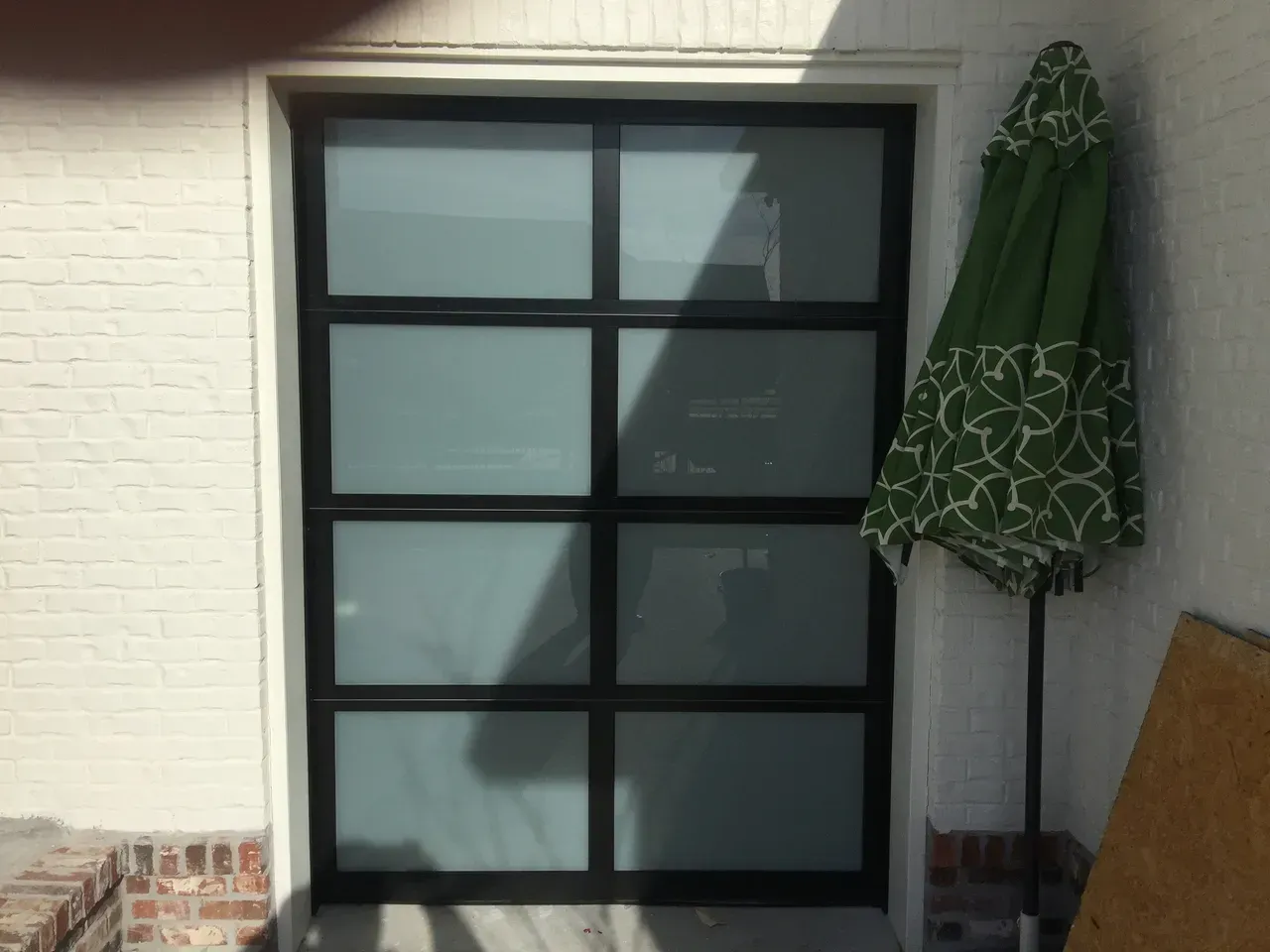 Black-framed glass door with four panes. Green patterned patio umbrella to the right. White brick wall.