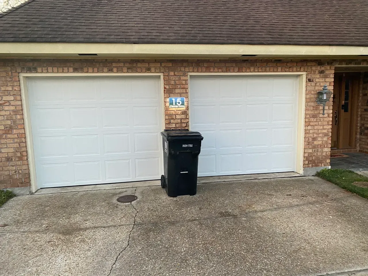 Two white garage doors with a black trash can in front. Brick building.