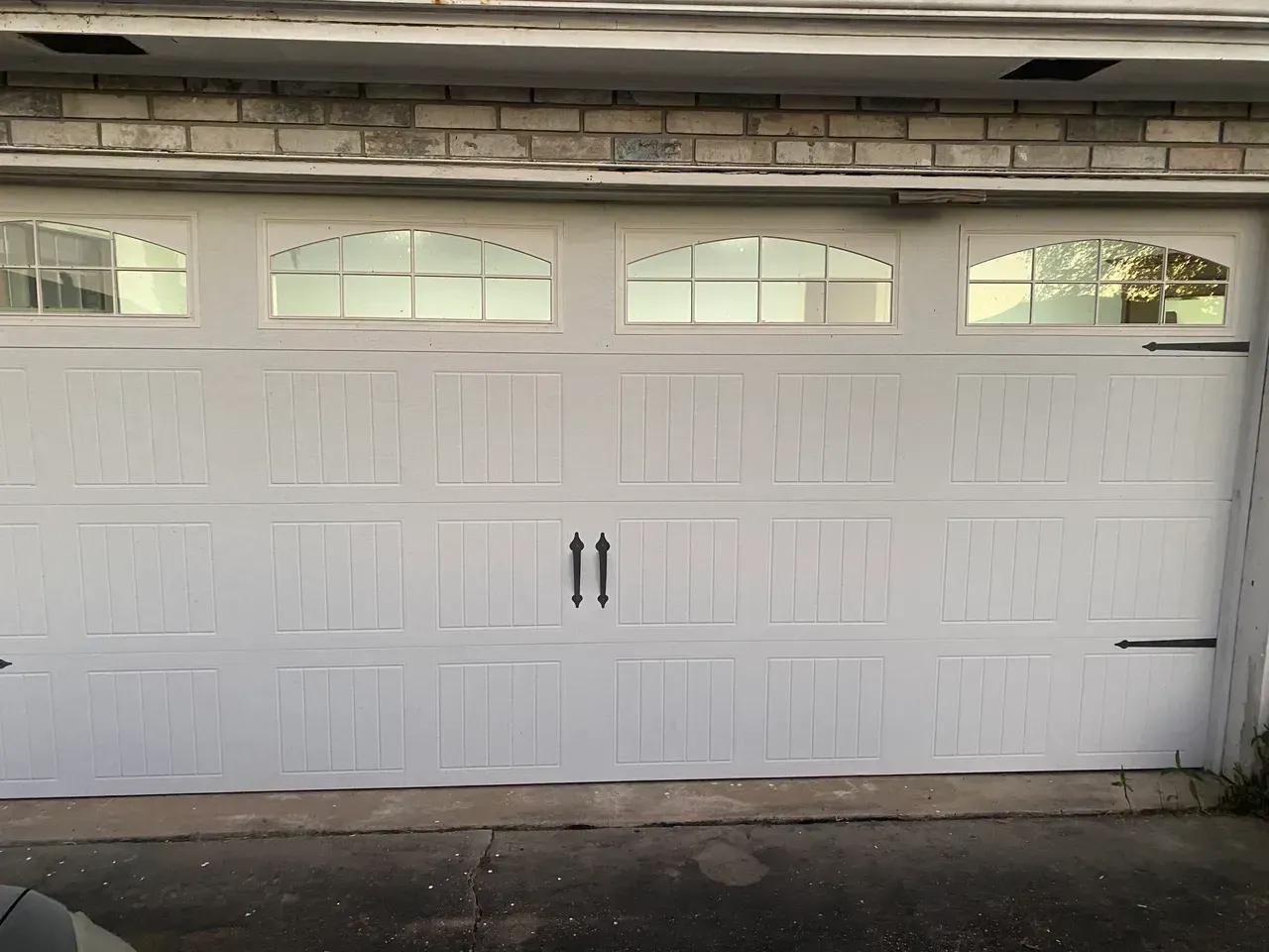 White garage door with arched glass panels above, black handles and hinges, against a brick wall.