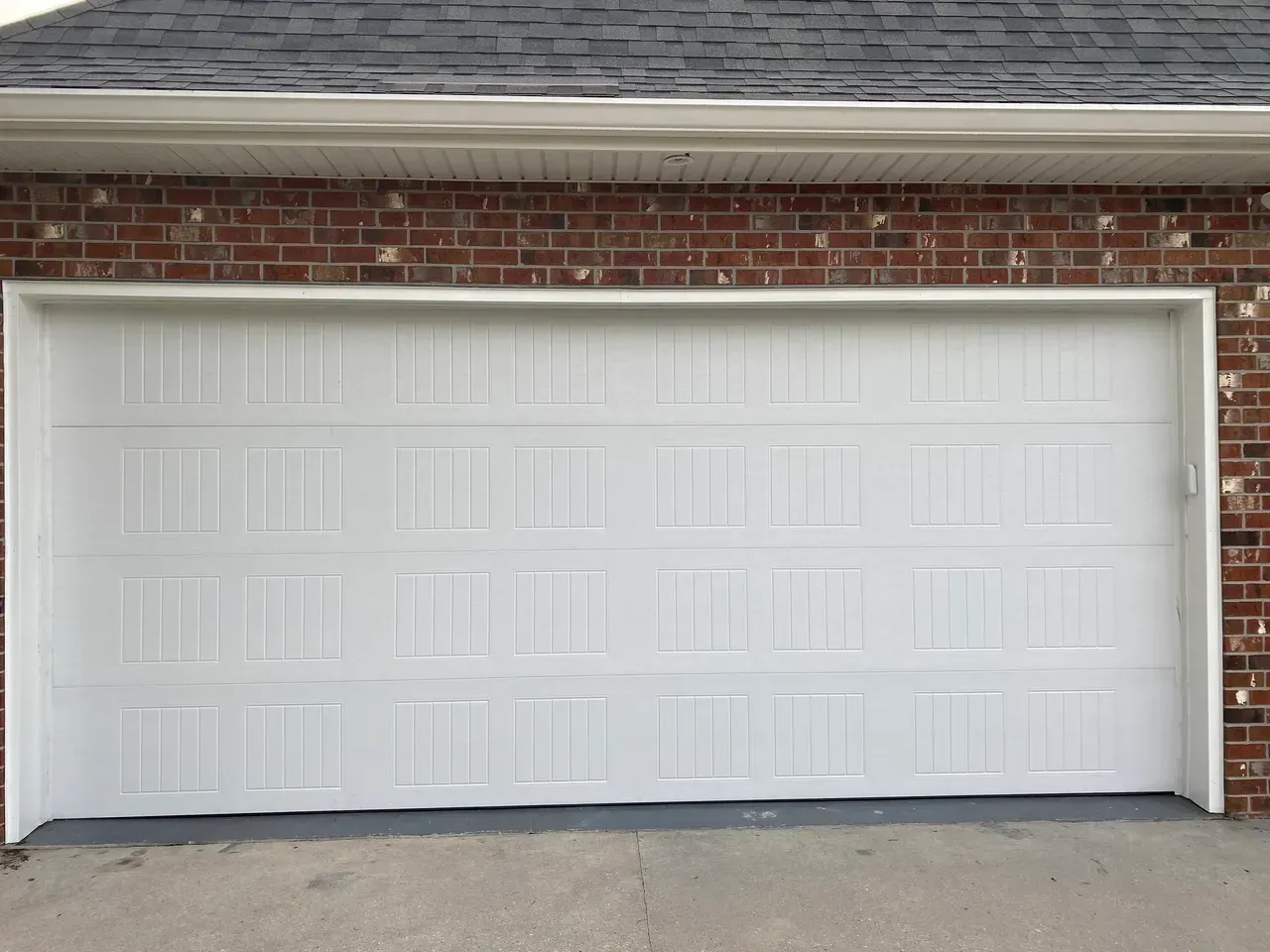White, paneled garage door set in a red brick building with a gray roof and concrete driveway.