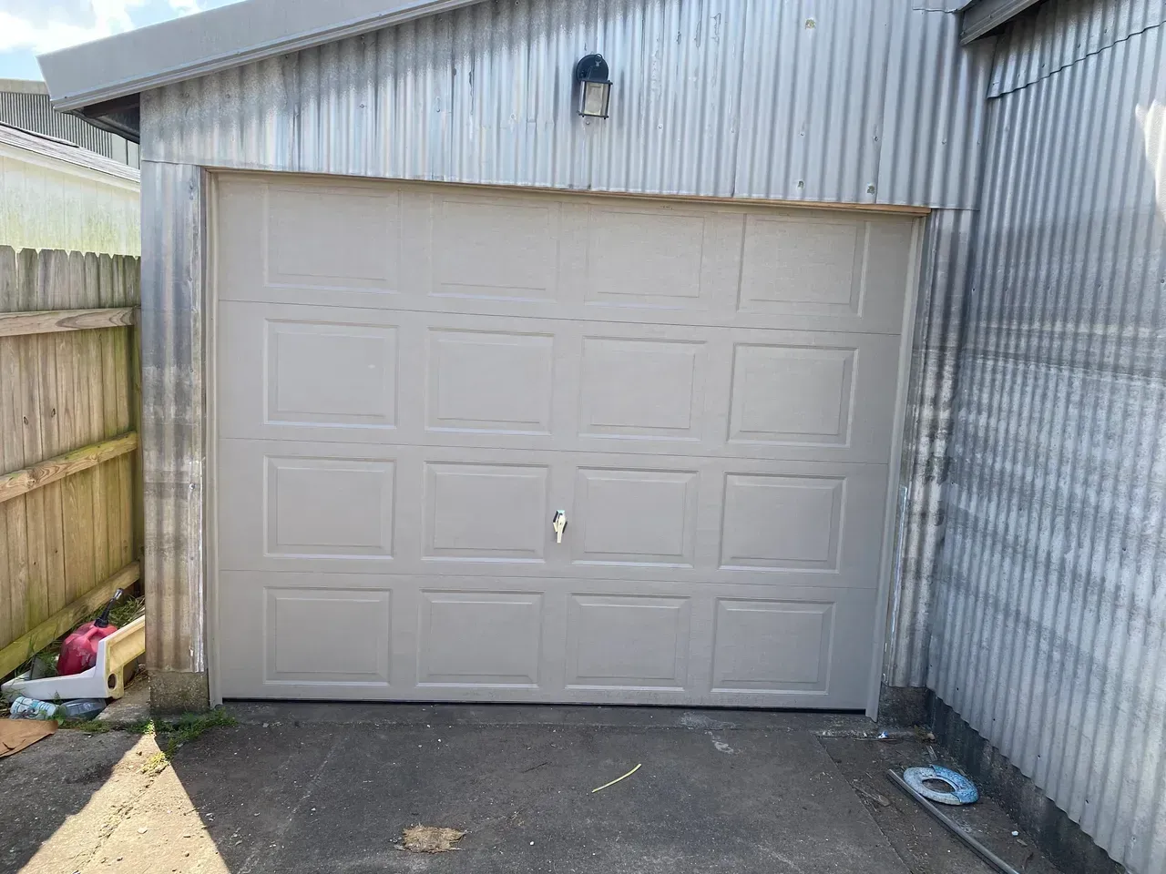 Tan garage door on a corrugated metal building with a weathered fence and ground.