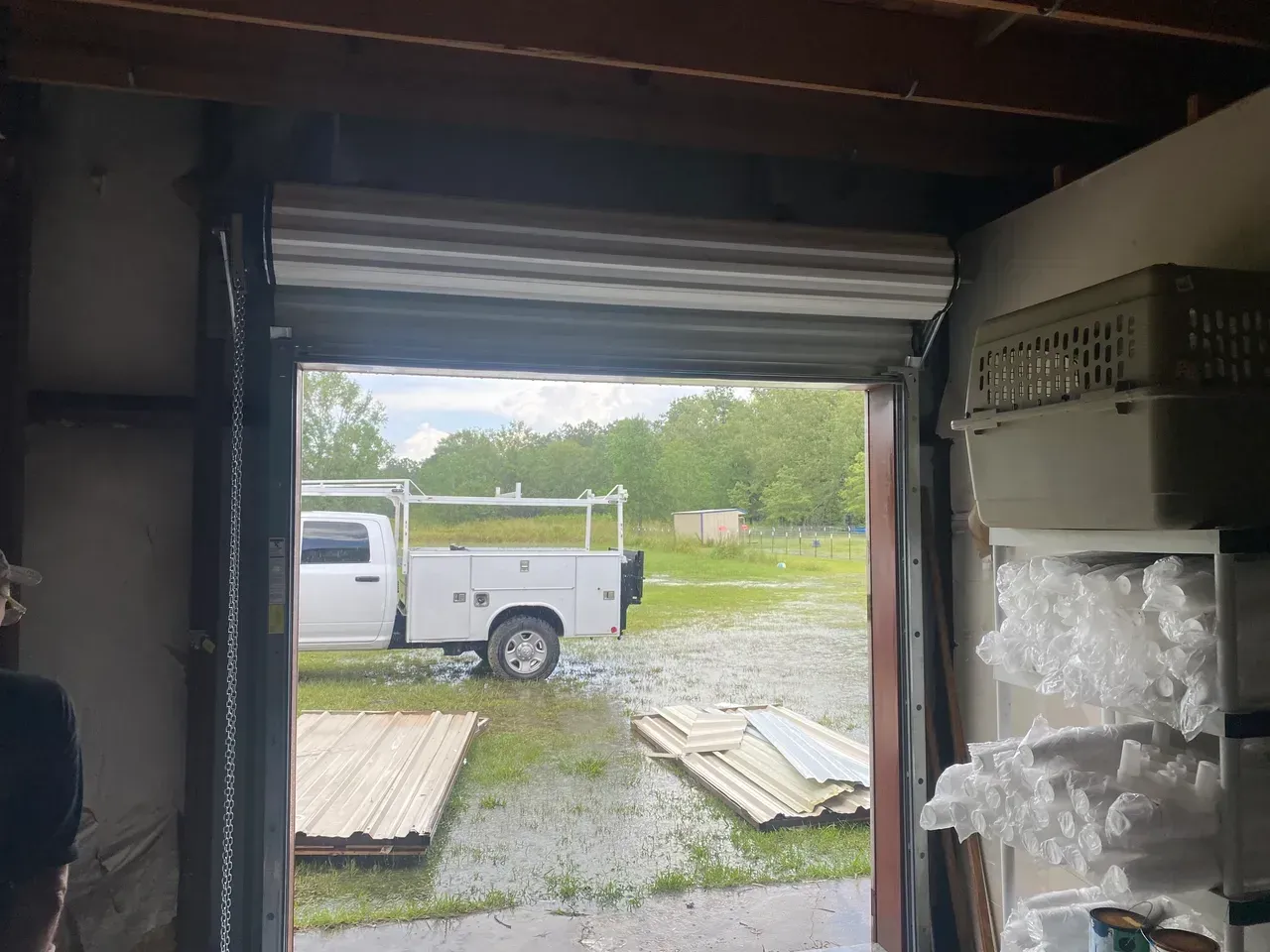 Open roll-up garage door frames a white truck parked outside. Overcast sky, flooded lawn.