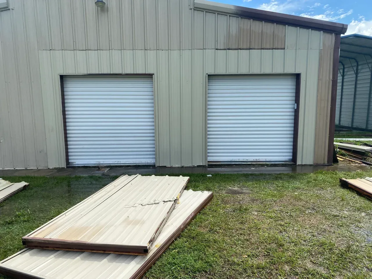Two closed garage doors on a light-colored building, with metal panels lying on the grass in front.