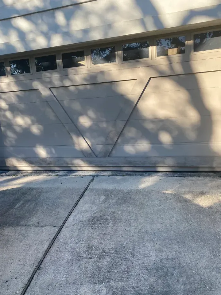 Tan garage door with triangle design, casting tree shadows on gray concrete driveway.