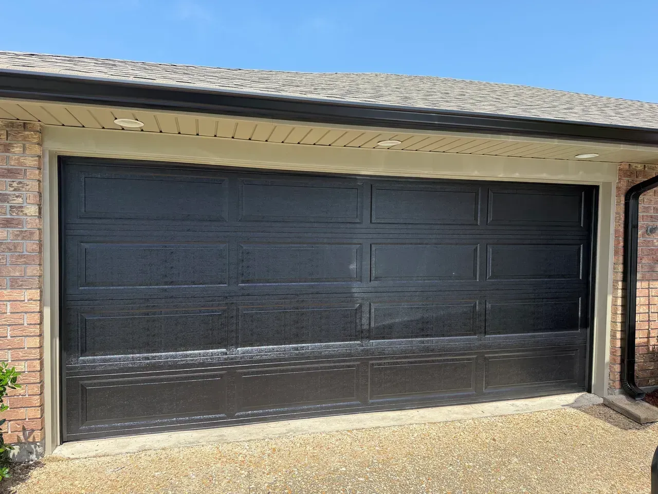 Black garage door on a brick building with a gravel driveway under a blue sky.