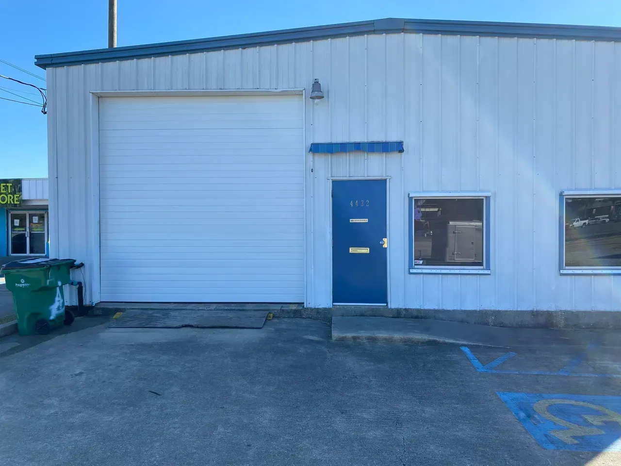 White building with a roll-up door, blue door, and windows. Green trash can and handicap parking space are present.