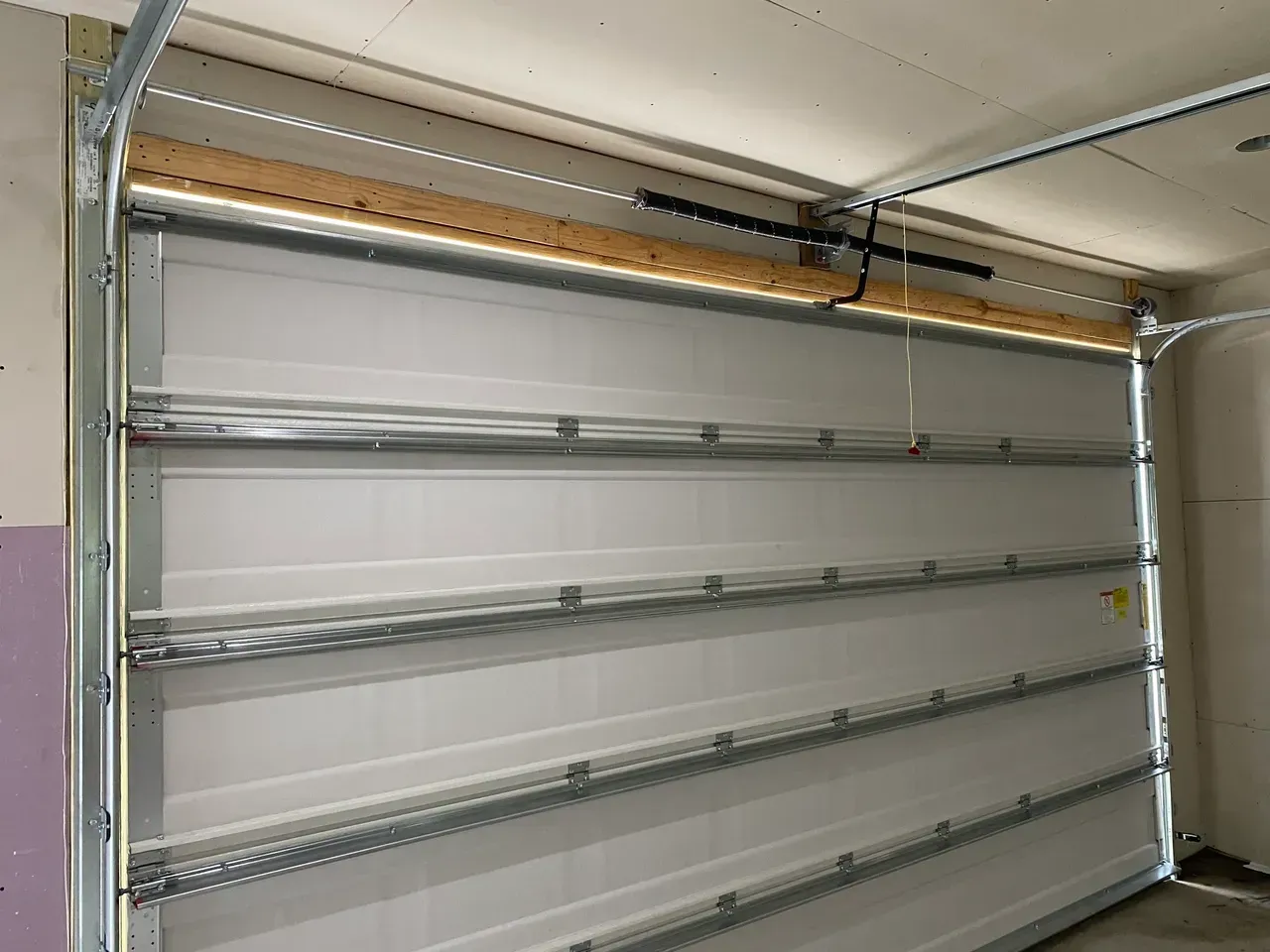 Garage door, partially open. White panels with metal framing, wooden header, and opener track visible.