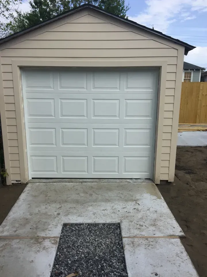White garage door on a light tan garage building with a concrete driveway and gravel square.