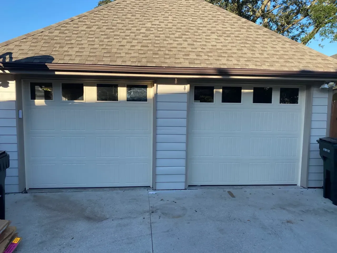Two beige garage doors with windows, under a brown roof. Concrete driveway.