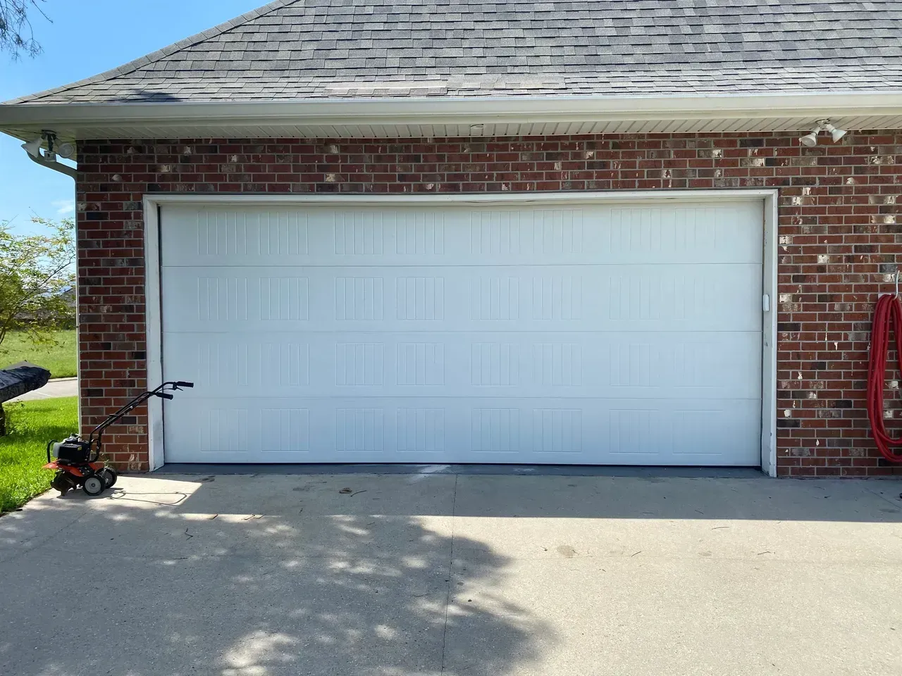 White garage door on brick building with a concrete driveway; a lawnmower is to the left, and a hose is to the right.