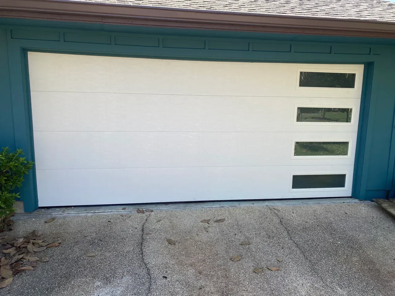 White garage door with three horizontal window panels, teal house exterior, concrete driveway.