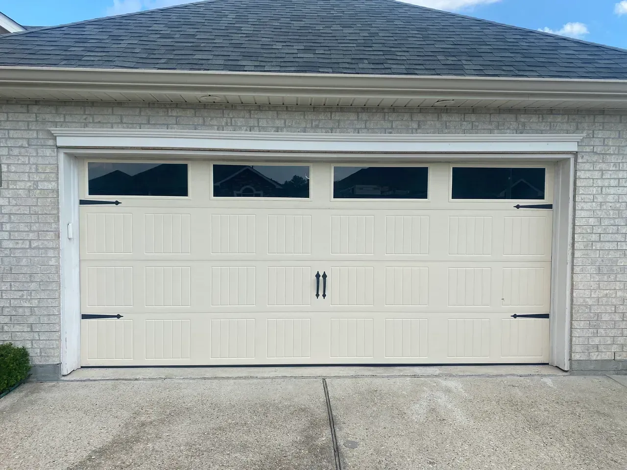 Cream-colored garage door with four small black-tinted windows, black hardware, and gray concrete driveway.