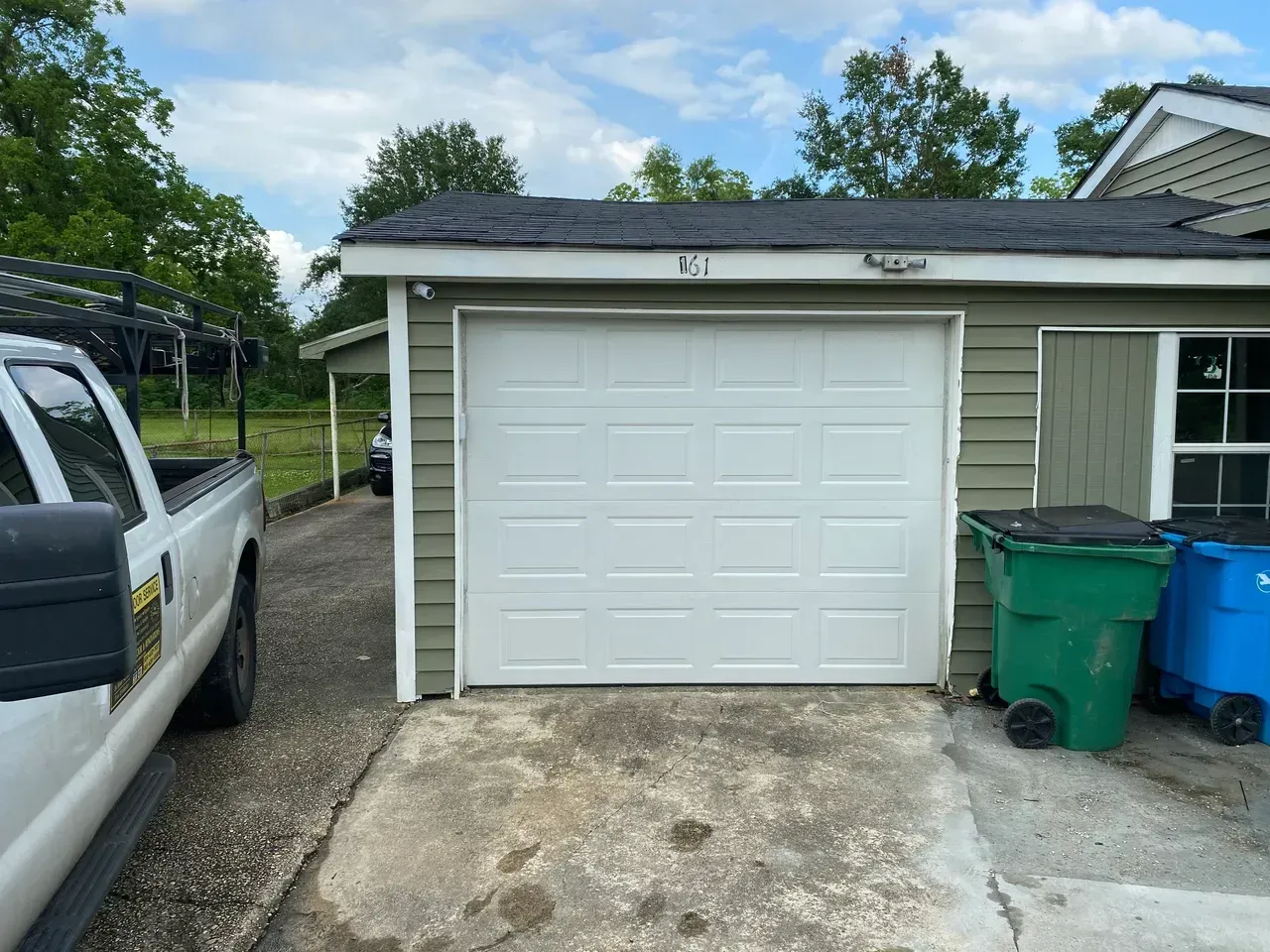 White garage door on a light green building with two trash cans next to it; a white truck is parked on the left.