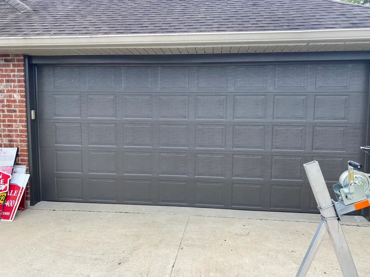 Gray garage door on a brick building; concrete driveway, and sawhorse visible.