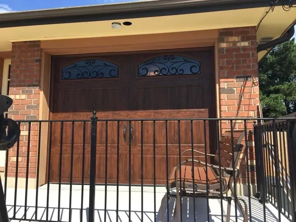 Brown garage door with decorative glass panes, enclosed by a black metal railing and brick columns.