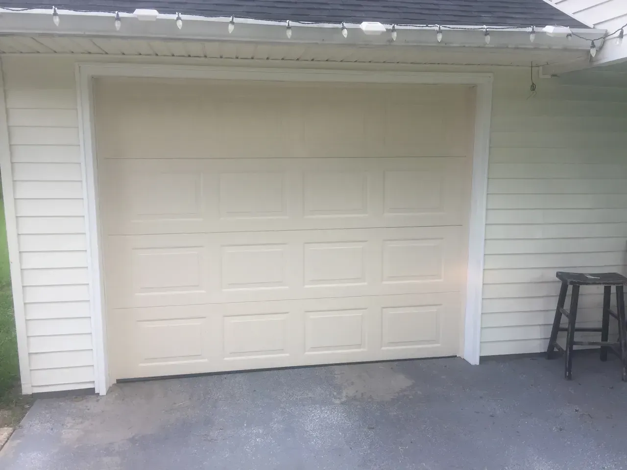 Tan closed garage door with white trim on a light beige siding building. A dark stool sits to the right.