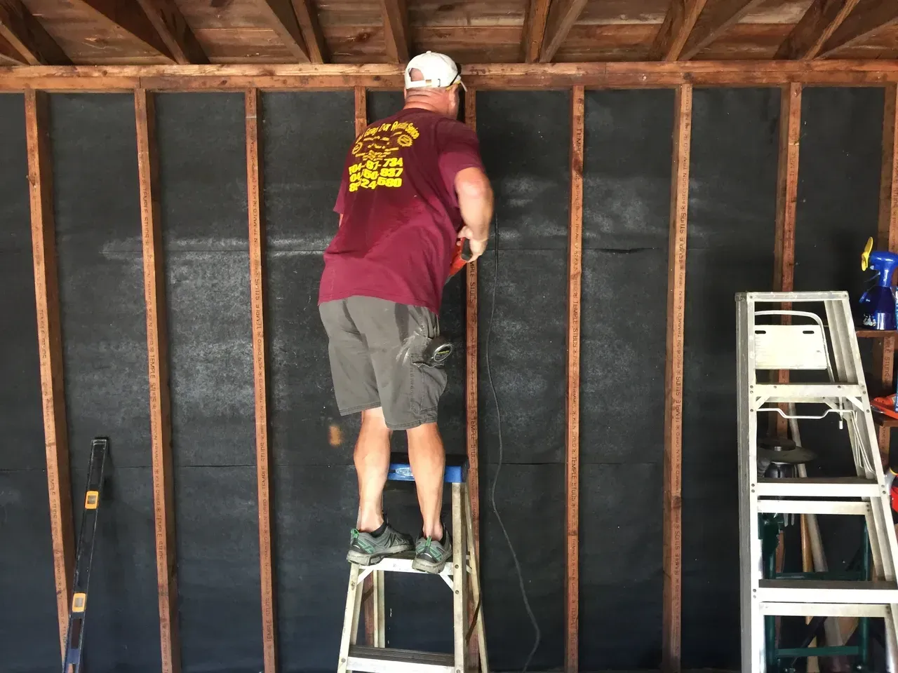 Man on stepladder cutting material on a garage wall. He's wearing a shirt, shorts, and a hat. Interior setting.