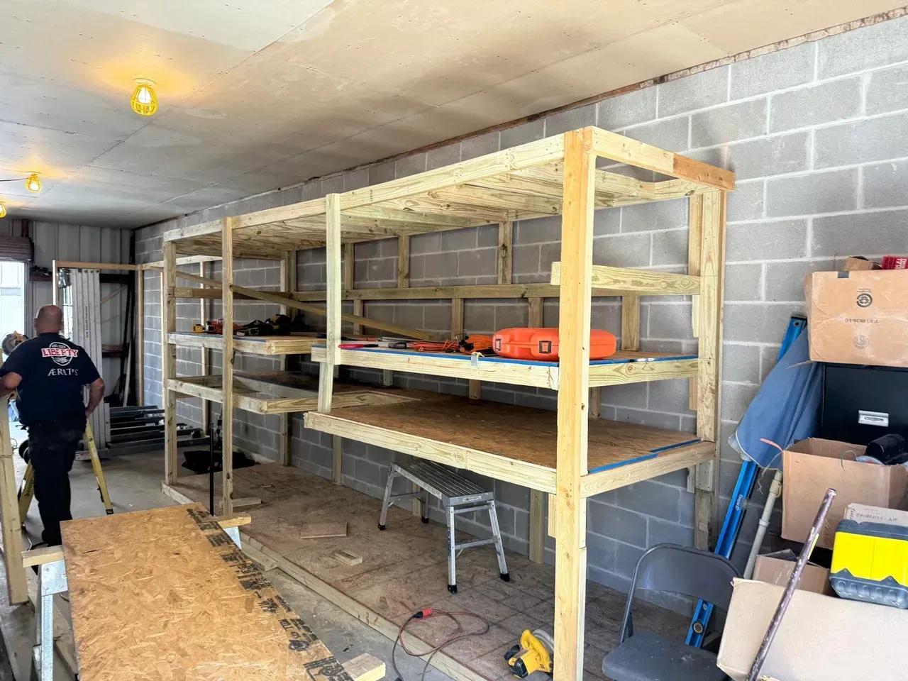 Wooden shelving unit against a cinder block wall, partially constructed, with tools and a person walking by.