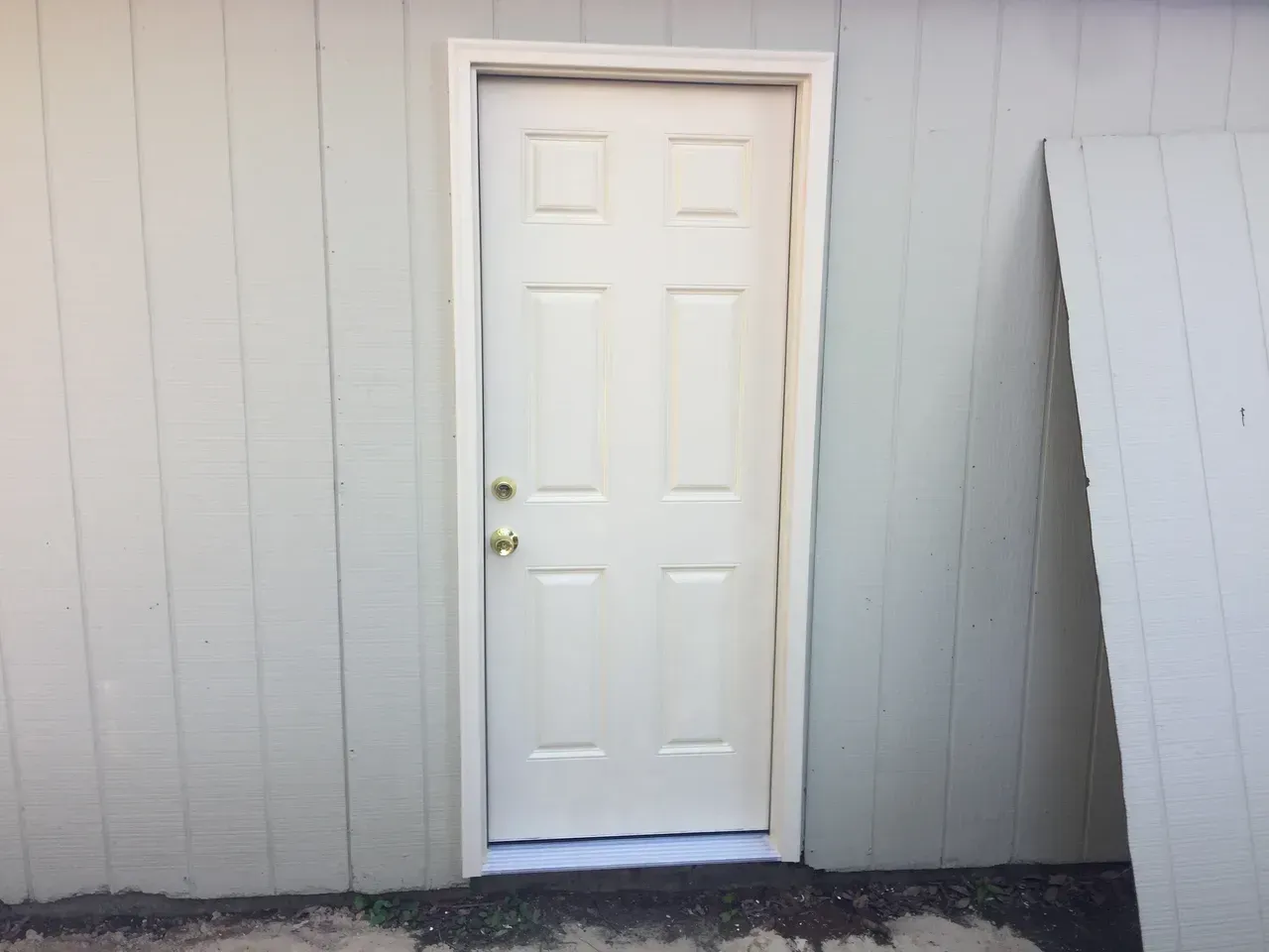 White door set in a light-colored building exterior, with trim and a partial wall panel leaning against it.