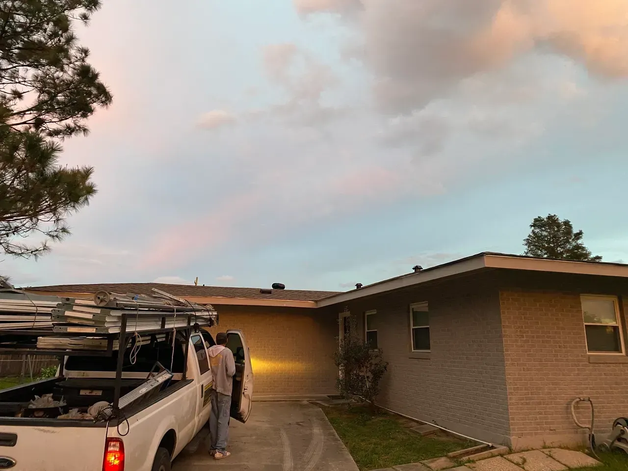 Person near a white pickup truck parked in a driveway in front of a house. Sunset sky.