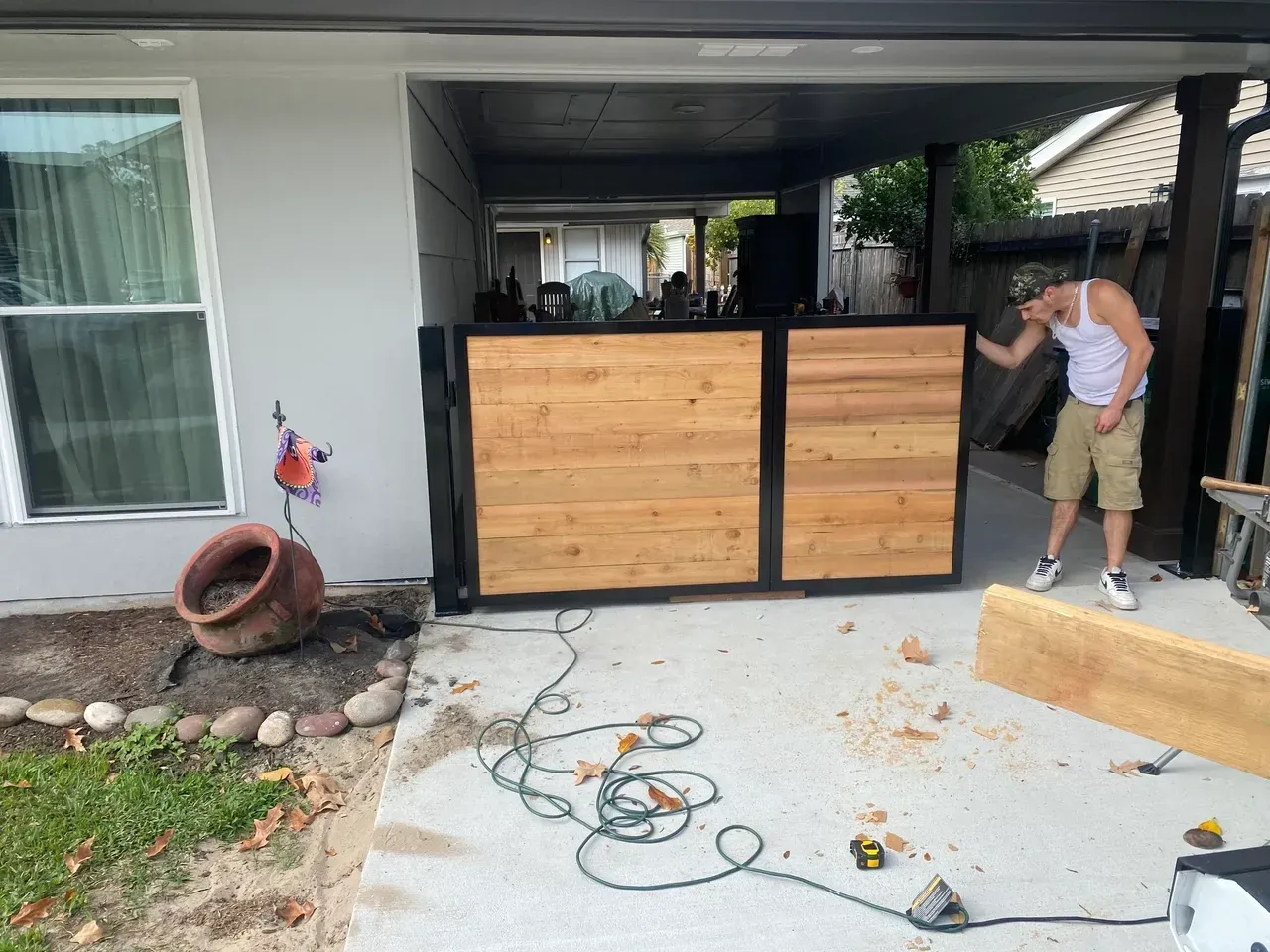 Man adjusts newly built wooden gate. Concrete patio, light gray building, and tools visible.