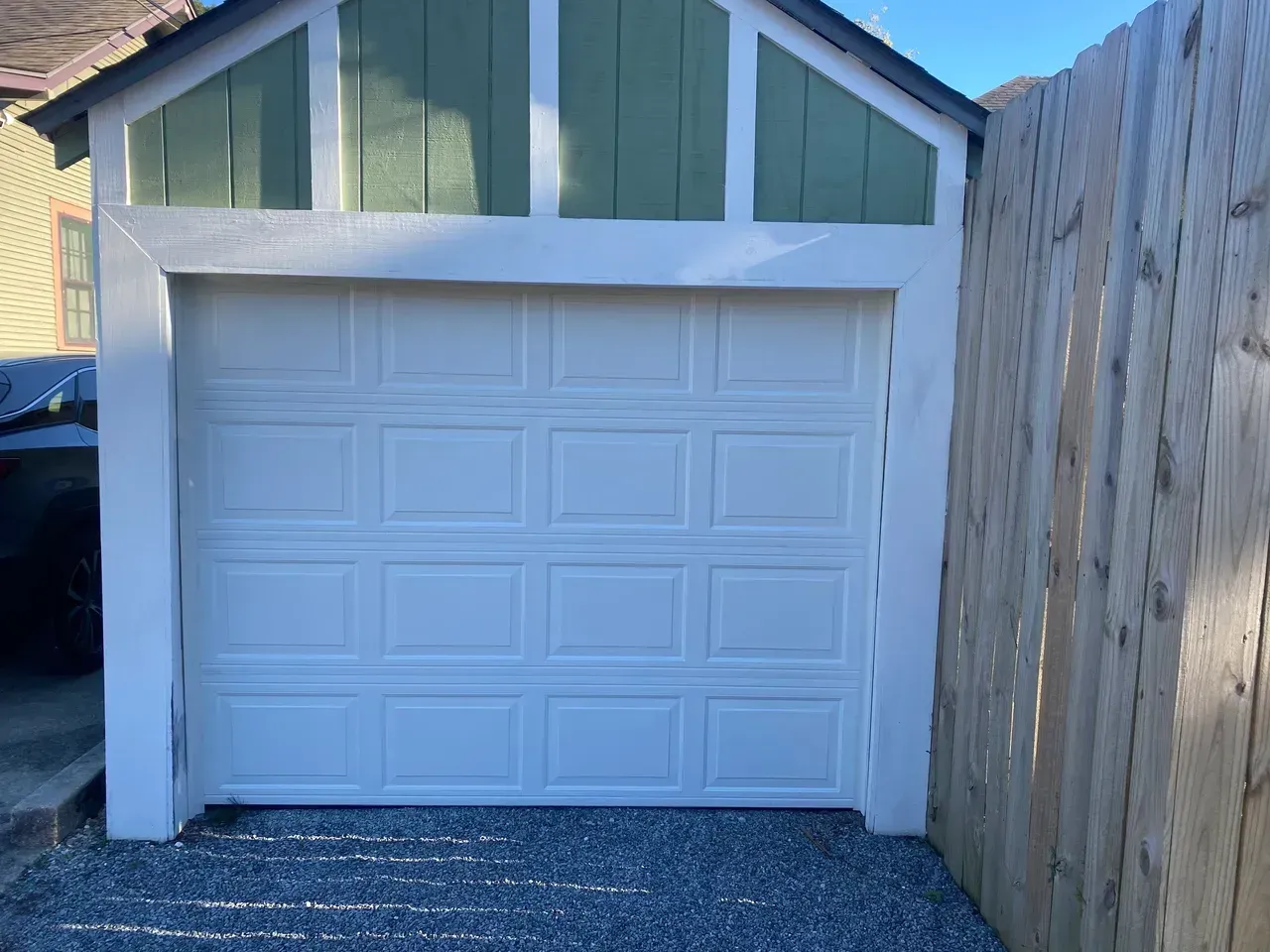 White garage door in a light-colored stucco frame. Gravel driveway. Green and white roof section. Wooden fence on the right.