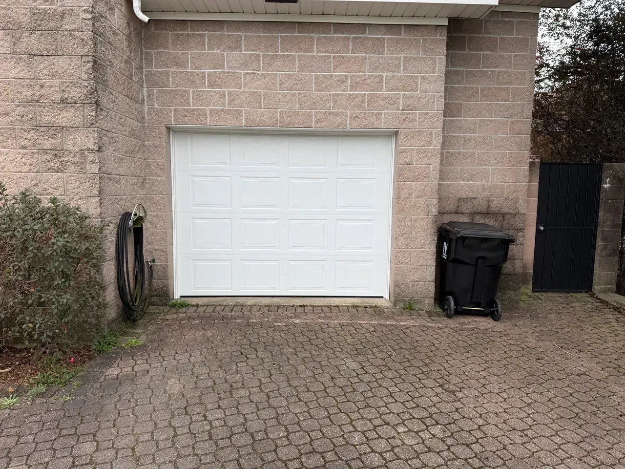 White garage door in a brick building with a black trash can and paved driveway.