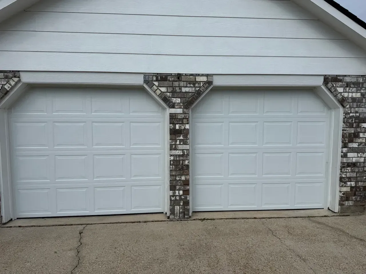 Two white garage doors with brick accents under a white house siding roof.