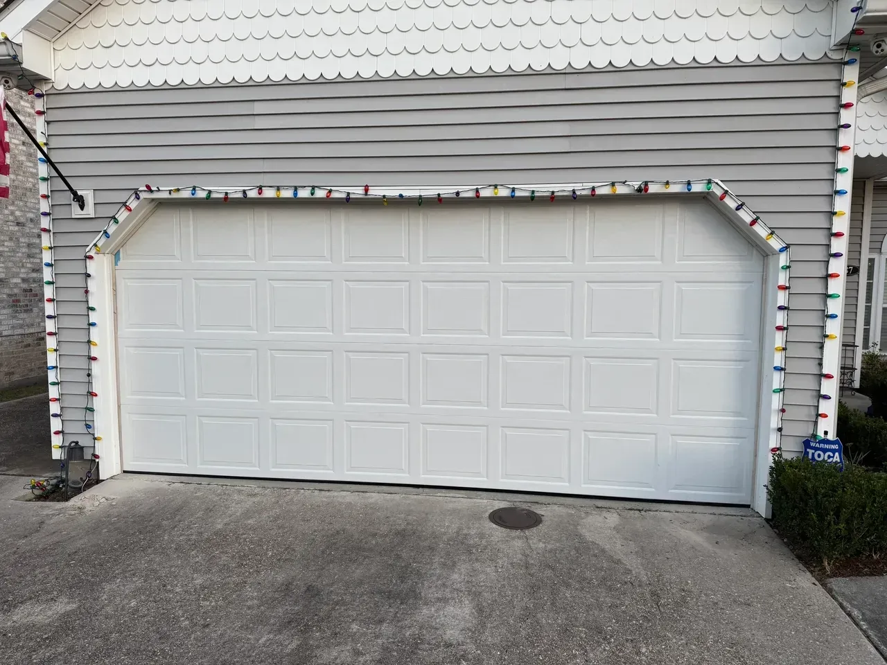 White garage door with Christmas lights on gray siding. Concrete driveway.