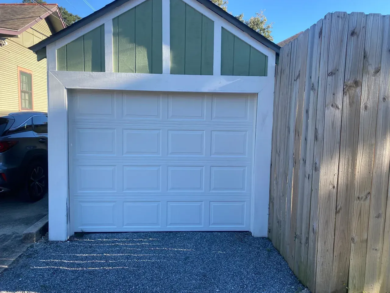 White garage door, green gabled roof, and gravel driveway next to a wooden fence.