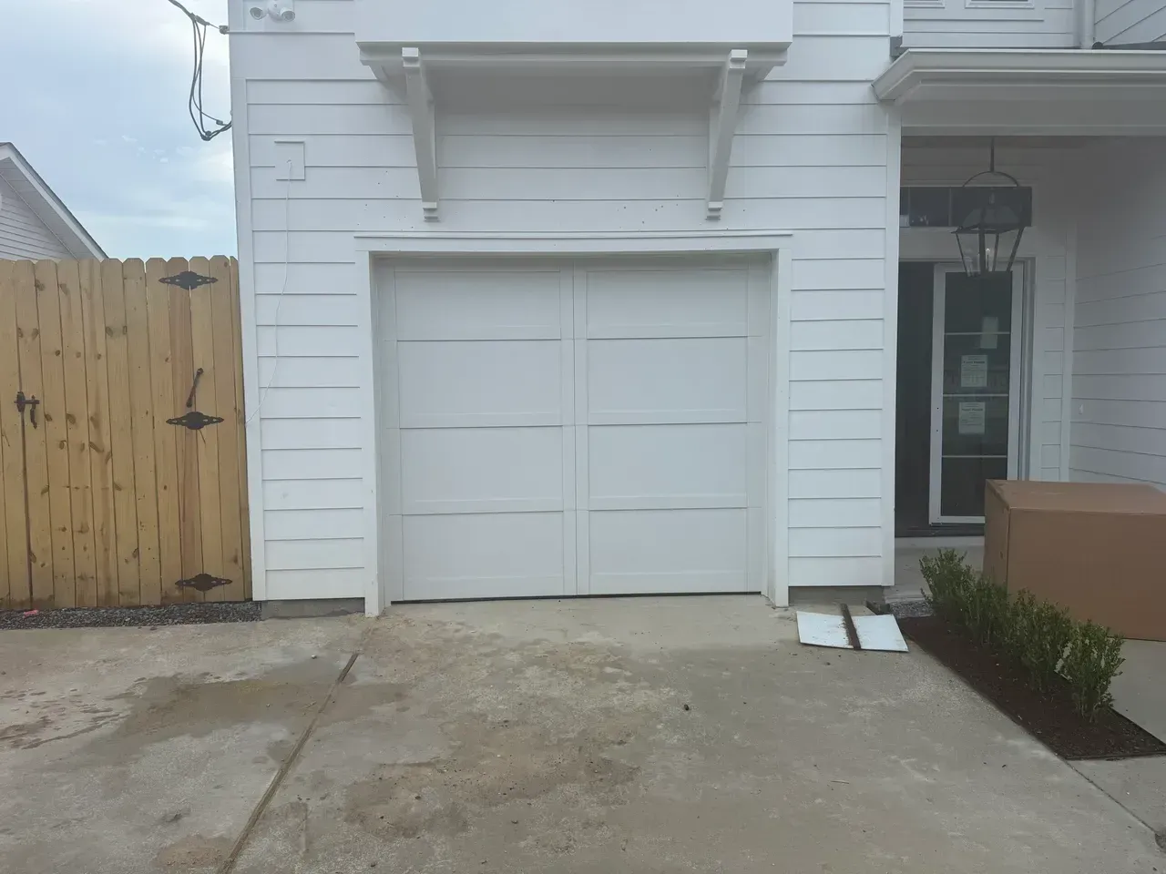 White garage door with concrete driveway, wooden fence on the left and a building with a door on the right.