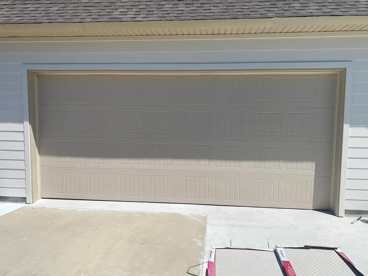 Beige garage door on a light-colored building, with a concrete driveway in front.