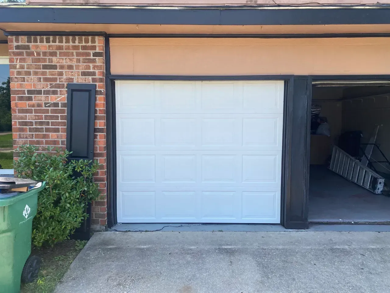 White garage door in a tan and brick building; a green trash can and greenery are visible.
