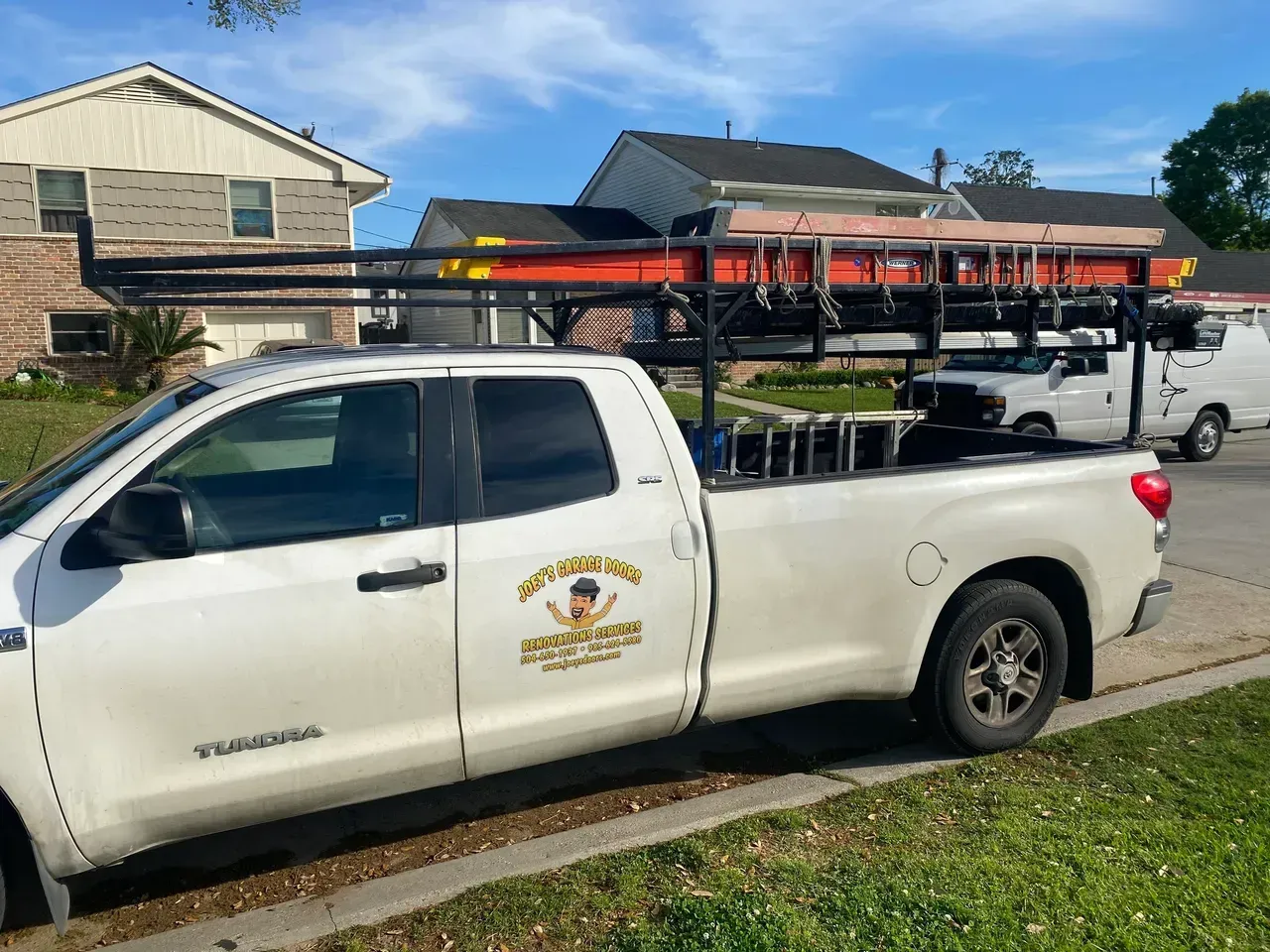White pickup truck with a ladder rack carrying ladders parked on a grassy verge.