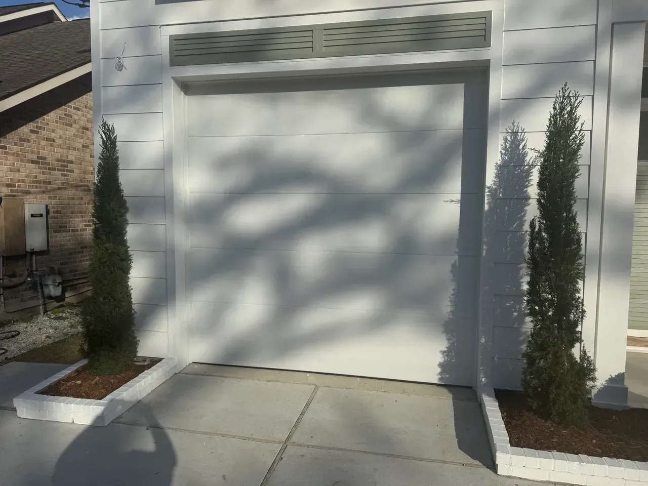 White garage door with two green trees in planters; concrete driveway.