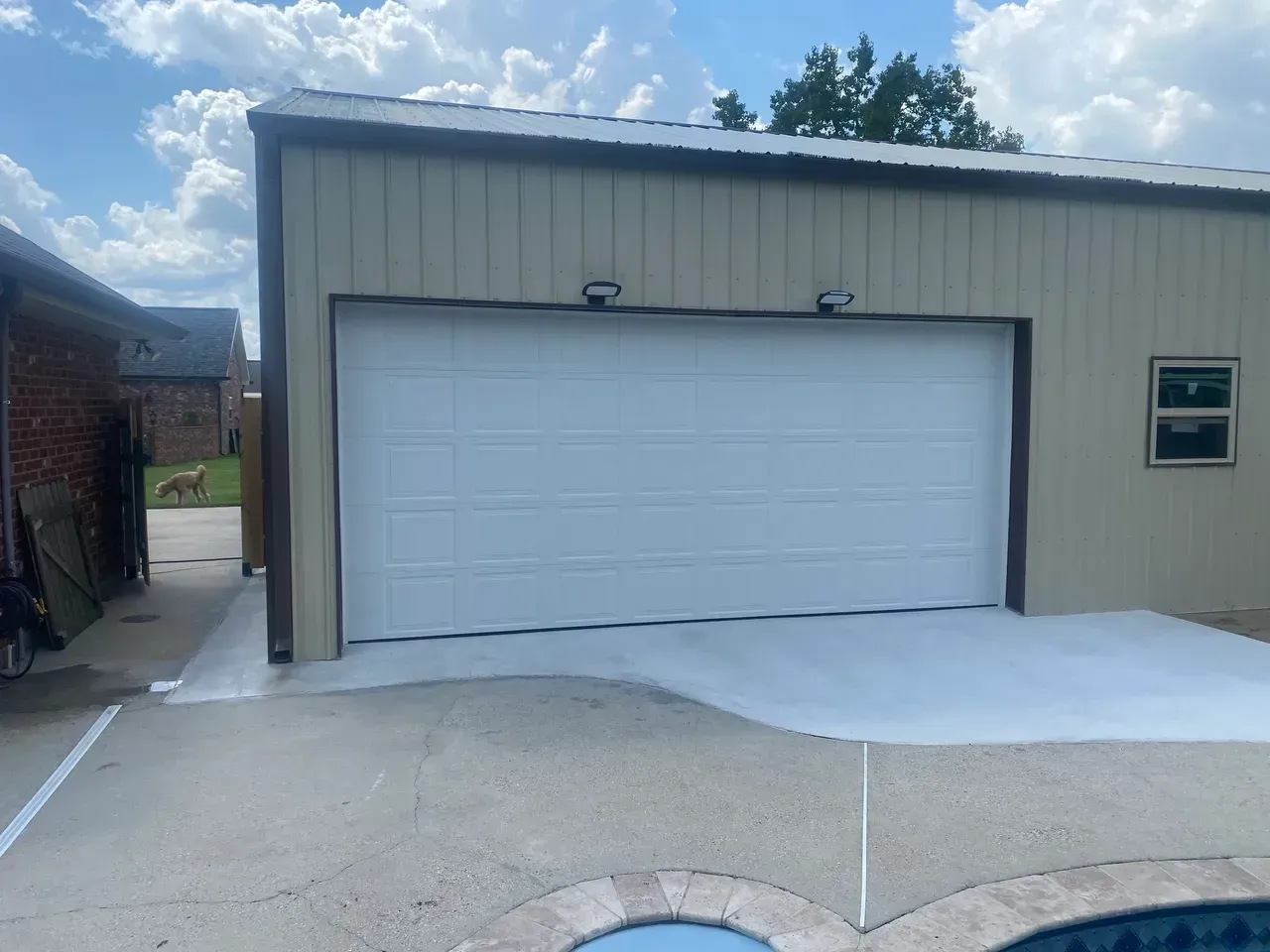 White garage door on tan metal building with concrete driveway and pool.