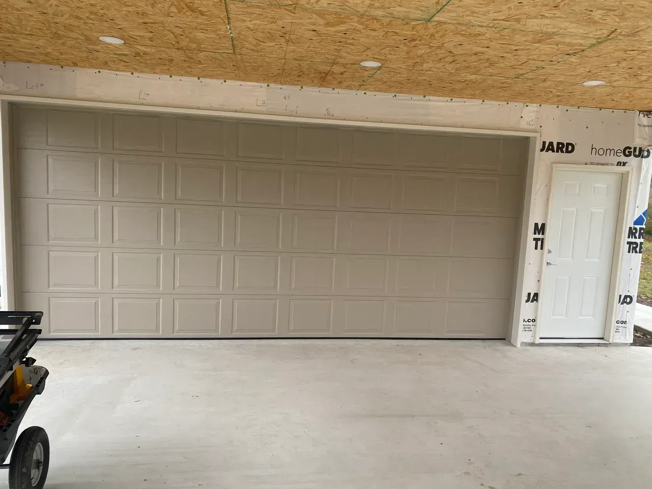 Tan garage door and white door on a building under construction, with a concrete floor.