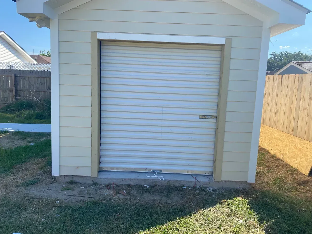 Tan shed with a closed, rolling garage door; set on concrete with beige siding and a grassy yard.