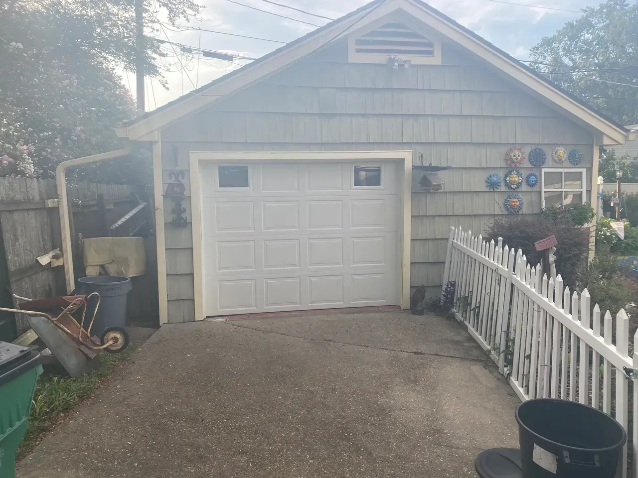 White garage with a gray driveway, bordered by a white picket fence and a wooden fence.