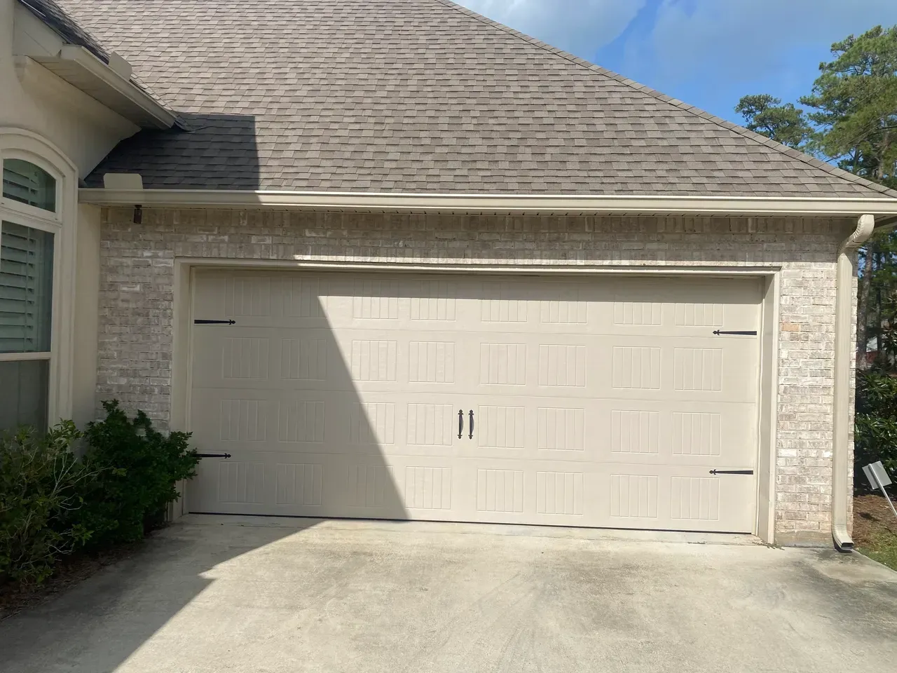 Beige garage door on a brick house with brown roof and concrete driveway.