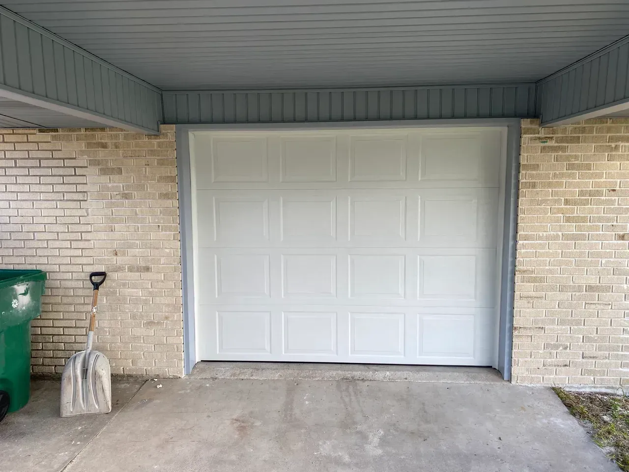 White garage door framed by gray trim, under a light brick awning. A green trash bin and shovel are on the left.