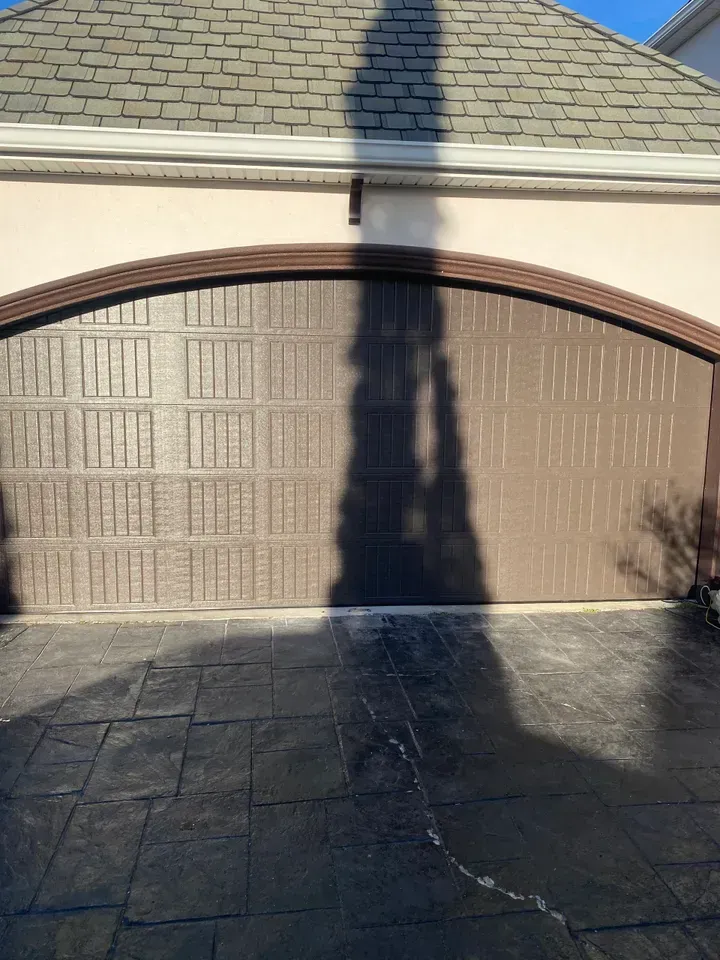 Brown arched garage door, shadow, beige house exterior, dark patterned driveway.