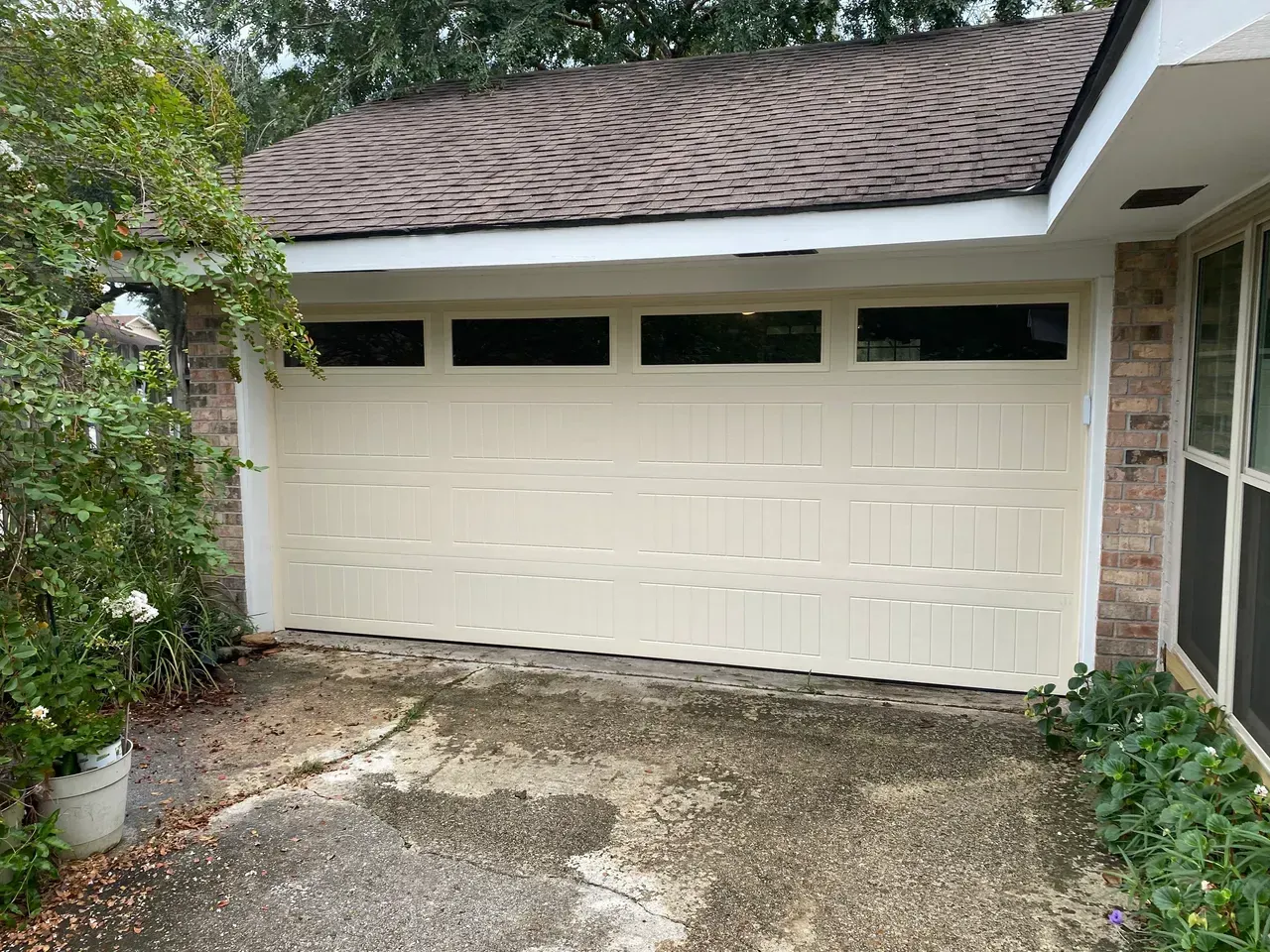 Tan garage door with windows, on a house with brown roof and brick exterior; overgrown greenery.