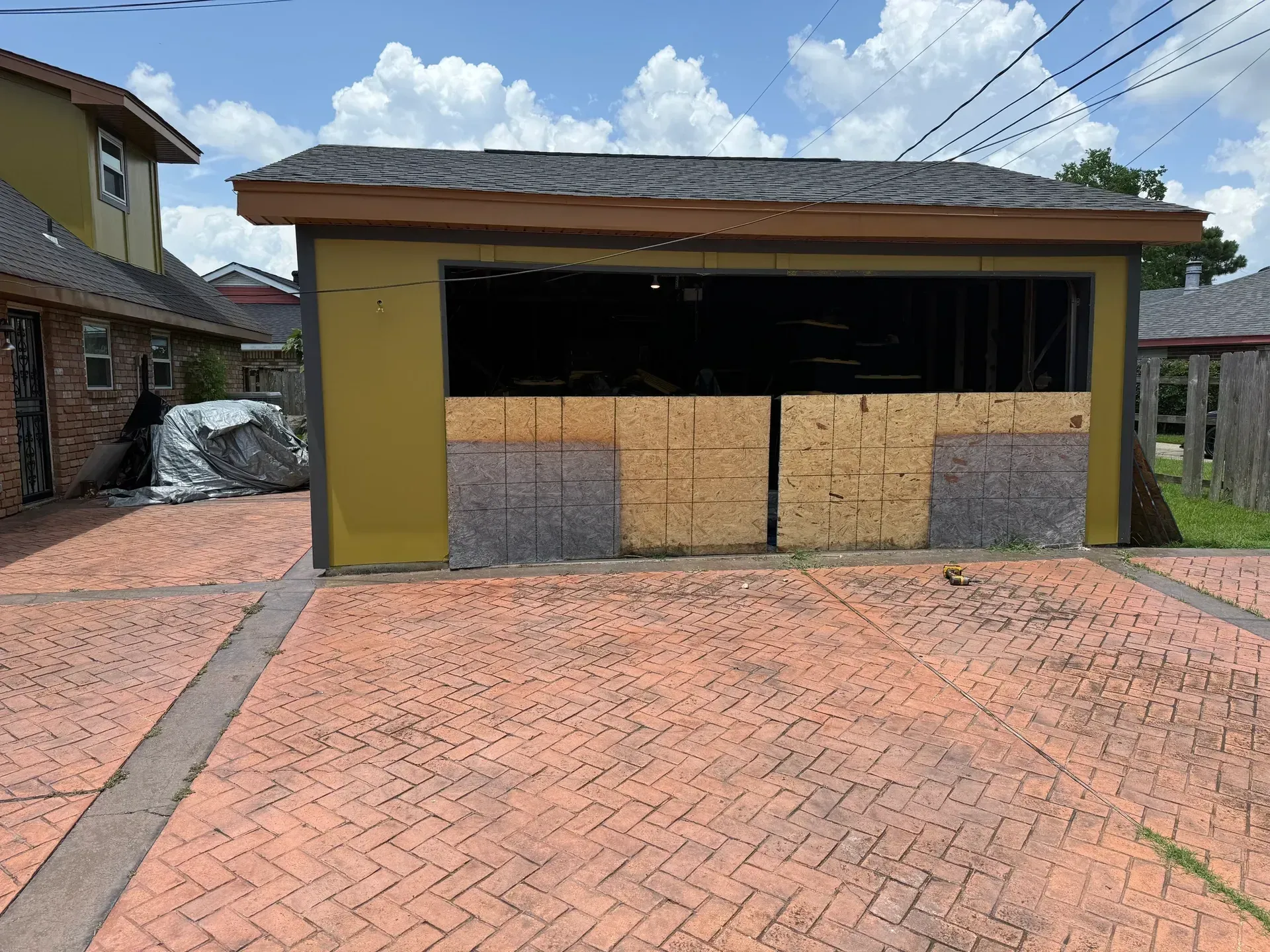 Garage with boarded-up openings, on a brick-patterned driveway. Yellow building with brown trim, under a cloudy sky.