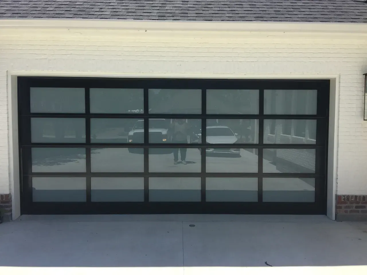 Black-framed glass garage door reflecting vehicles and person, set in a white-walled building with a concrete driveway.