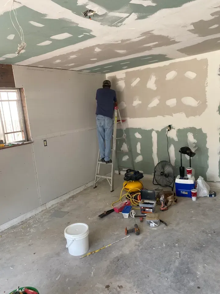 Person on a ladder installing drywall in a room with unfinished walls and ceiling. Tools and supplies on the floor.