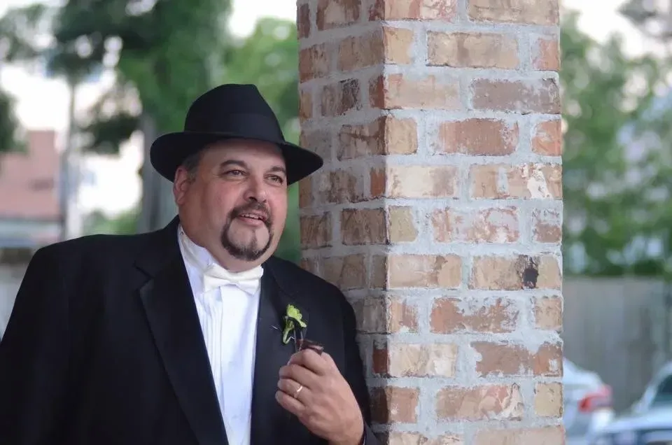 Man in tuxedo and fedora leans against a brick pillar, smiling.