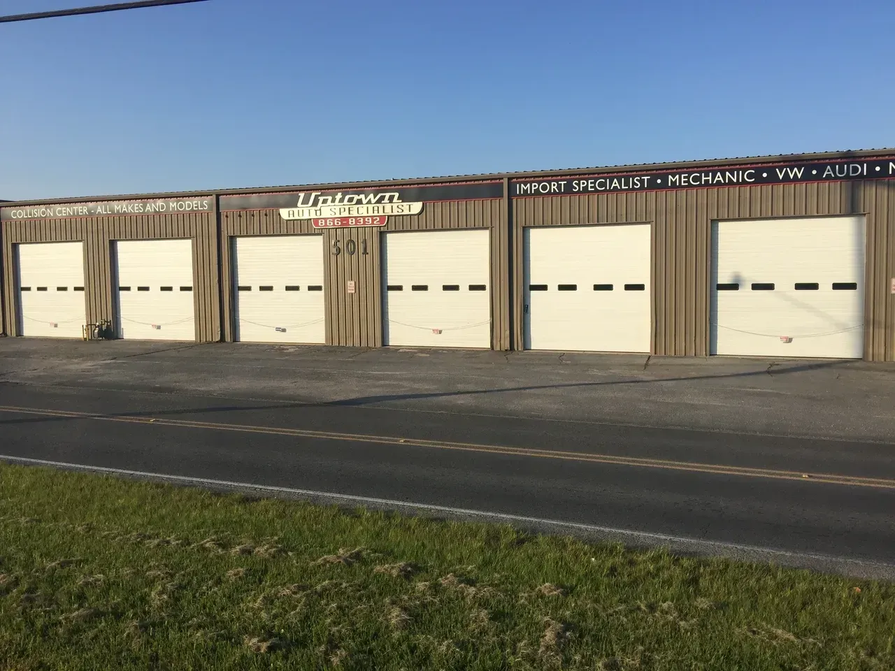 Exterior view of a repair shop with five closed garage doors, a sign, and a road in front.