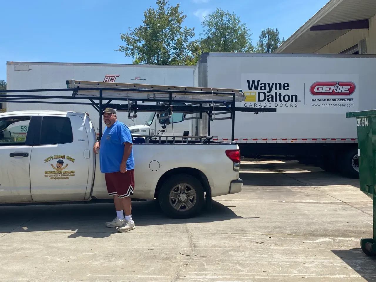 Man standing by a white pickup truck parked next to two large white trucks. Sunny day.