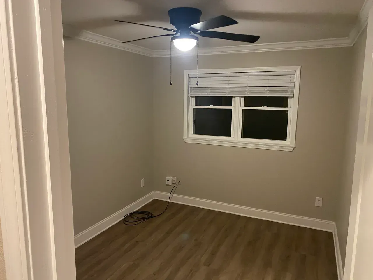 Empty room with tan walls, brown flooring, and a window with blinds. A ceiling fan with a light is on.